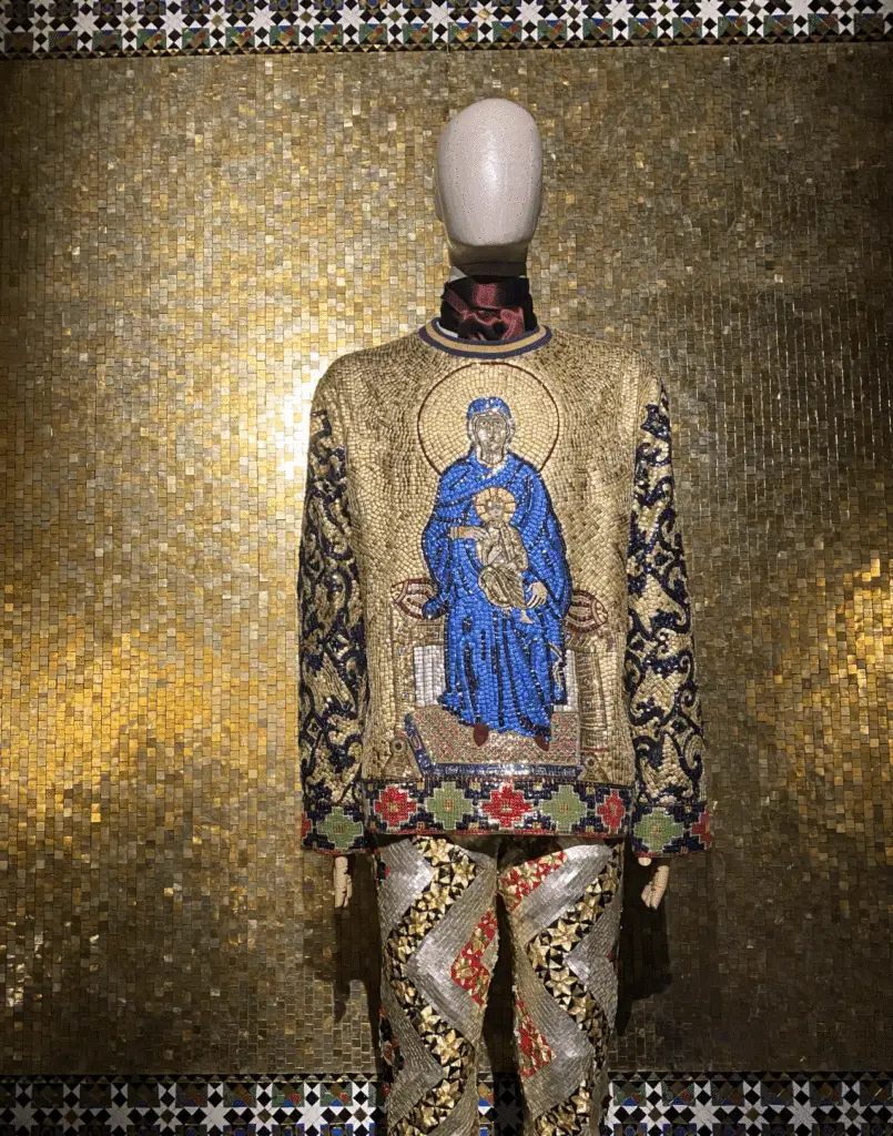 Mannequin in ornate gold and jeweled D&G outfit featuring a Byzantine-style Madonna and Child motif, set against a golden mosaic wall.