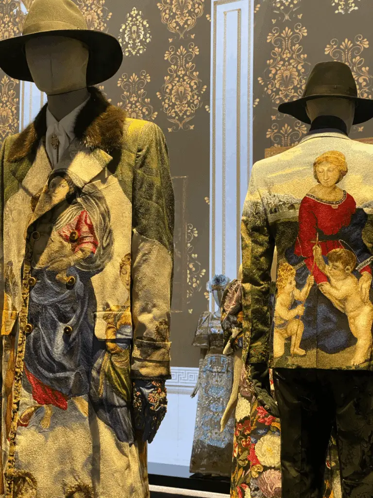 Two mannequins in embroidered D&G coats with Renaissance-style religious and mythological scenes, accessorized with wide-brim hats