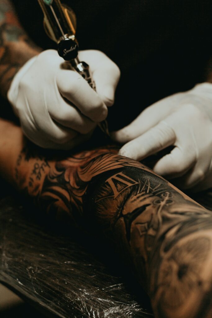 A tattoo artist wearing white gloves carefully inks a detailed black and gray sleeve tattoo on a client’s arm using a tattoo machine.