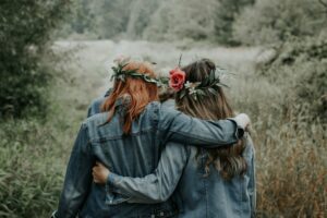 Two friends walking arm in arm outdoors wearing flower crowns, representing the Galentine’s Day cultural movement and friendship