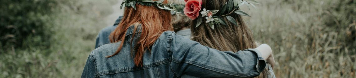Two friends walking arm in arm outdoors wearing flower crowns, representing the Galentine’s Day cultural movement and friendship
